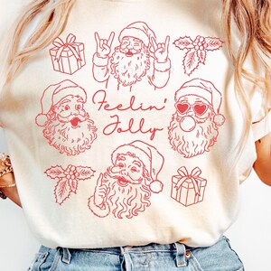 May include: Cream-colored t-shirt featuring red illustrations of Santa Claus with various expressions, gifts, and holly. The phrase "Feelin' Jolly" is written in red script. The shirt is made of a soft material.