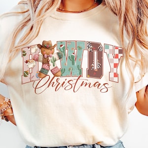Może przedstawiać: Kremowa koszulka z napisem "Howdy Christmas" w stylu western. Słowo "Howdy" ma wzór patchworku, kaktusy i nadruk krowy. Piernikowy kowboj znajduje się na środku. Słowo "Christmas" jest napisane kursywą.
