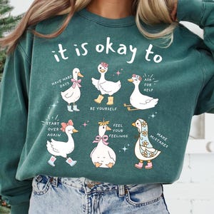 Pode incluir: Camisola verde-azulado com a frase "it is okay to" acima de gansos de desenho animado com mensagens encorajadoras como "Have hard days" e "Make mistakes". Os gansos usam acess&oacute;rios como botas e la&ccedil;os.
