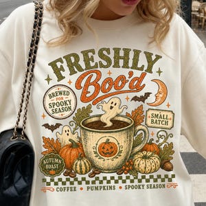 Könnte beinhalten: Ein cremefarbenes Sweatshirt mit einem Grafikdruck mit dem Text FRESHLY Boo'd, BREWED FOR SPOOKY SEASON, SMALL BATCH, AUTUMN ROAST, COFFEE, PUMPKINS, SPOOKY SEASON. Das Design zeigt eine Kaffeetasse, Kürbisse, Geister und einen Halbmond.