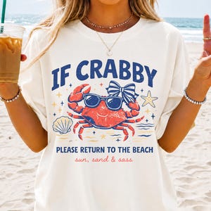May include: Cream-colored t-shirt with the text "IF CRABBY PLEASE RETURN TO THE BEACH" and a graphic of a crab wearing sunglasses. The design includes a bow, starfish, and seashell. The words "sun, sand & sass" are also included.