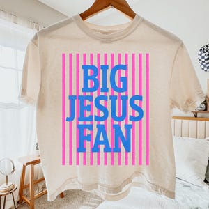 May include: Cream-colored t-shirt with the text "BIG JESUS FAN" in blue block letters, set against a background of pink vertical stripes. The shirt is hanging on a wooden hanger.