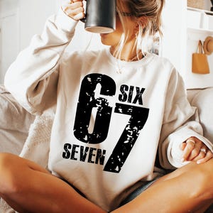 May include: Cream-coloured sweatshirt featuring the number "67" in a distressed black font. The words "SIX" and "SEVEN" are above and below the number. A black mug is held up near the face.