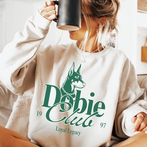 Può includere: Felpa color crema con grafica verde di un Dobermann e le parole "Dobie Club" con "Loyal Legacy" sotto. La felpa ha maniche lunghe e girocollo. Una tazza nera è tenuta davanti al viso.