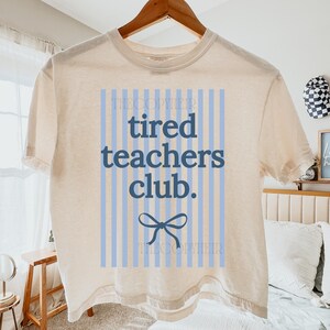 May include: Cream-colored t-shirt with the text "tired teachers club." in blue, set against a light blue striped background. A blue bow graphic is below the text. The shirt is hanging on a wooden hanger.