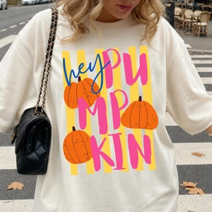 May include: Cream-colored t-shirt with the text "hey PUMPKIN" in blue, pink, and yellow. The design includes orange pumpkin illustrations. The shirt is a casual, oversized fit.