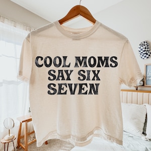 May include: Cream-colored t-shirt with the text "COOL MOMS SAY SIX SEVEN" in bold, black letters. The shirt is hanging on a wooden hanger. The style is casual and relaxed, suitable for everyday wear.