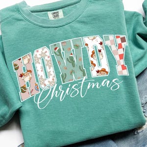 May include: A teal Comfort Colors sweatshirt with the words "HOWDY Christmas" in a decorative font. The letters are filled with Christmas-themed patterns, including cacti, Santa hats, and snowflakes. The sweatshirt is folded on a denim surface.