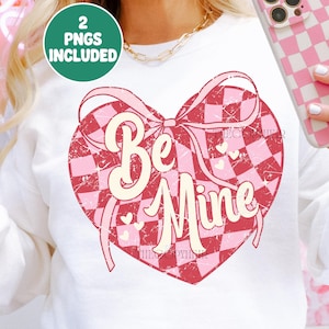 Puede incluir: Sudadera blanca con un corazón a cuadros rosa y rojo, un lazo y las palabras "Be Mine". El diseño incluye pequeños corazones y el texto "2 PNGS INCLUDED" en un círculo verde.