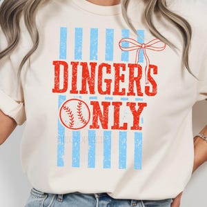 May include: Cream-colored t-shirt with the text "DINGERS ONLY" in red, a baseball graphic, and blue vertical stripes. A red bow is at the top right. The shirt is a casual style.