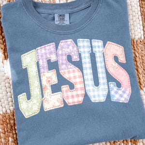 Può includere: Una maglietta blu Comfort Colors con la scritta "JESUS" applicata sul davanti. Le lettere sono realizzate con vari tessuti fantasia, tra cui floreali, quadretti e colori uniti. La maglietta è esposta su una superficie tessuta.