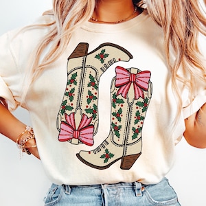 May include: Cream-colored sweatshirt featuring a graphic of two cowboy boots. The boots are decorated with holly and red berries, and each has a pink bow. The boots have brown heels and are set against a cream background.