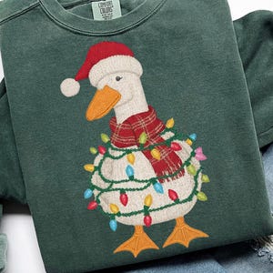 May include: Green Comfort Colors sweatshirt featuring an embroidered goose wearing a Santa hat, a red plaid scarf, and wrapped in Christmas lights. The goose has an orange beak and feet. The sweatshirt has a crew neck.