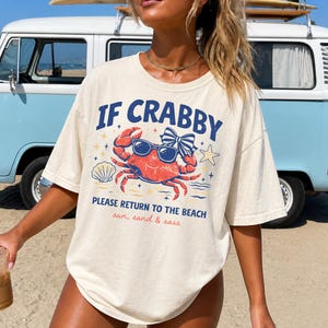 May include: Cream-colored t-shirt with a graphic of a crab wearing sunglasses and a bow. The text reads "IF CRABBY, PLEASE RETURN TO THE BEACH, sun, sand & sass." A vintage blue and white van is in the background.
