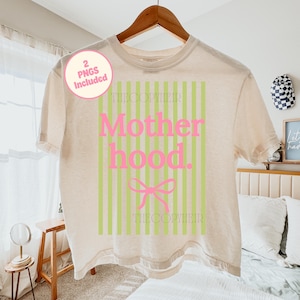 Puede incluir: Una camiseta color crema con la palabra "Motherhood" en rosa, sobre un fondo de rayas verticales verdes. Una cinta rosa está debajo de la palabra "hood". El texto "THECOPYHEIR" está impreso arriba y abajo del diseño. Un círculo rosa con el texto "2 PNGS Included" está en la parte superior izquierda.