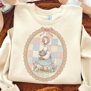 Puede incluir: Sudadera color crema con una ilustraci&oacute;n de un ganso con gorro y delantal, sosteniendo una cesta de huevos de Pascua. El ganso est&aacute; enmarcado en un &oacute;valo con un fondo de retazos. El texto "THE COPIER" es visible.