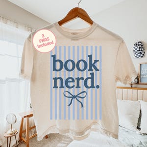 May include: A cream-coloured t-shirt with the words "book nerd" in dark blue, set against a light blue striped background. A blue bow graphic is below the text. The shirt is hanging on a wooden hanger.