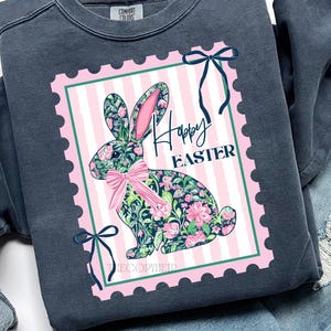 May include: A dark grey sweatshirt with a pink and white striped postage stamp design. The stamp features a floral bunny and the words "Happy Easter." The bunny has pink ears and a pink bow. Green bows are at the corners.