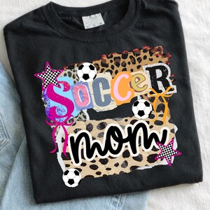 May include: Black t-shirt with a colorful graphic that says "Soccer Mom." The design includes soccer balls, stars, and a leopard print background. The word "Soccer" is in a collage style with different colors.