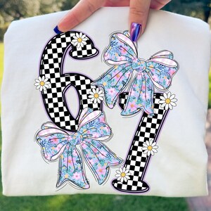 May include: Cream-colored t-shirt featuring the numbers "67" with a checkerboard pattern and floral bows. The numbers are outlined in black and purple, with white daisies scattered around them. The bows have a floral pattern with pink, blue, and yellow flowers.