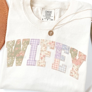 May include: Cream-colored t-shirt with the word "WIFEY" in patchwork letters. The letters feature floral and gingham patterns in pastel colors. A gold necklace with a small pendant rests on the shirt.