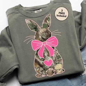 May include: A sage green sweatshirt featuring a bunny design with a camouflage pattern, a pink bow, and a heart. The image includes the text "3 PNGs Included" on a circular sticker.
