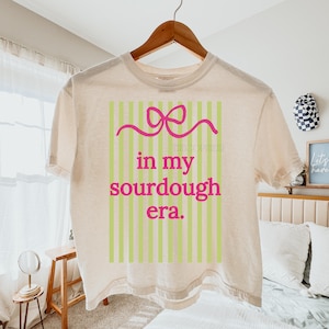 May include: A cream-colored t-shirt with a pink bow and the text "in my sourdough era" in a pink font. The text is set against a background of vertical green stripes. The shirt is hanging on a wooden hanger.