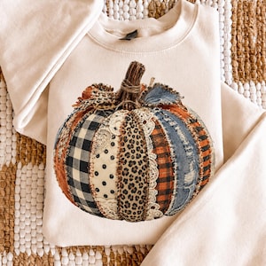 Può includere: Una felpa girocollo color crema con una grande stampa centrale raffigurante una zucca composta da vari ritagli di tessuto. Il design include segmenti con motivo a quadri bianchi e neri, pois, stampa leopardata, denim e inserti in pizzo.