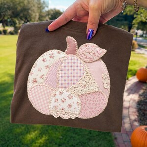 May include: A dark brown t-shirt featuring a large pumpkin applique made from various fabric patches. The pumpkin design includes light pink and white floral, gingham, and plaid patterns, accented with cream-colored lace trim and stitching.