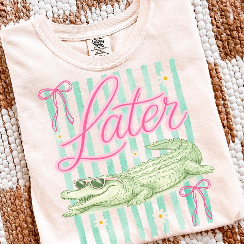 K&ouml;nnte beinhalten: Hellpfirsichfarbenes T-Shirt mit Grafikdesign. Das Design zeigt das Wort "Later" in rosa Schrift, ein gr&uuml;nes Alligator mit Sonnenbrille und rosa Schleifen. Der Hintergrund hat hellblaue Streifen und kleine wei&szlig;e Blumen.