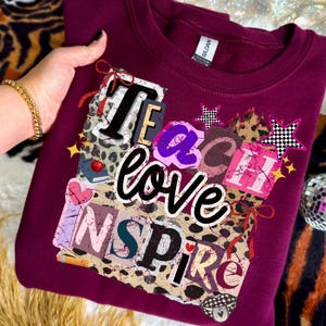 May include: A maroon sweatshirt with the words "Teach Love Inspire" in a collage design. The design includes leopard print, stars, hearts, and ribbons. The sweatshirt is a Gildan brand.