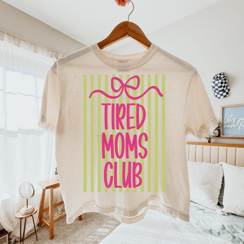 K&ouml;nnte beinhalten: Ein cremefarbenes T-Shirt mit dem Text "TIRED MOMS CLUB" in Pink, vor einem Hintergrund aus gr&uuml;nen und wei&szlig;en vertikalen Streifen. Ein rosa Schleifendesign befindet sich &uuml;ber dem Text. Das Shirt h&auml;ngt an einem Holzb&uuml;gel.