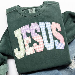 Puede incluir: Sudadera verde oscuro con la palabra "JESUS" aplicada en la parte delantera. Las letras est&aacute;n hechas de varias telas estampadas, incluyendo flores, cuadros y colores lisos. La sudadera tiene cuello redondo y mangas largas.