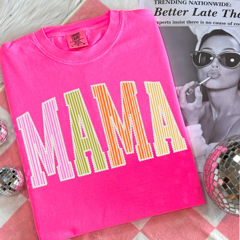 Puede incluir: Una camiseta rosa brillante con la palabra "MAMA" en letras grandes y rayadas multicolores. Las letras est&aacute;n delineadas con un borde blanco festoneado. La camiseta est&aacute; sobre una superficie a cuadros rosa y blanco.