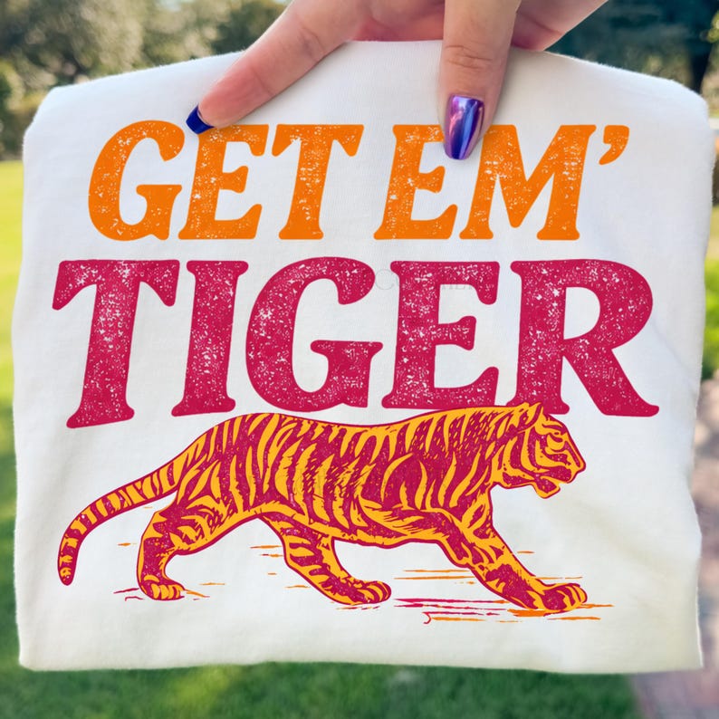 K&ouml;nnte beinhalten: Wei&szlig;es T-Shirt mit dem Text "GET EM' TIGER" in Orange und Rot, mit einer Tiger-Illustration in Gelb und Rot. Das Design ist auf der Vorderseite des Shirts aufgedruckt.