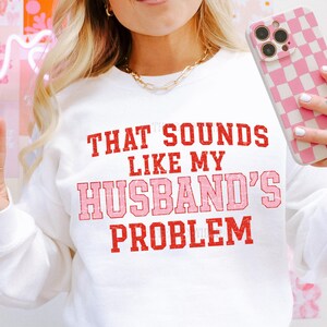 May include: White sweatshirt with the text "THAT SOUNDS LIKE MY HUSBAND'S PROBLEM" in red and pink lettering. The sweatshirt is being held by a person wearing a gold chain necklace. A pink and white checkered phone case is also visible.