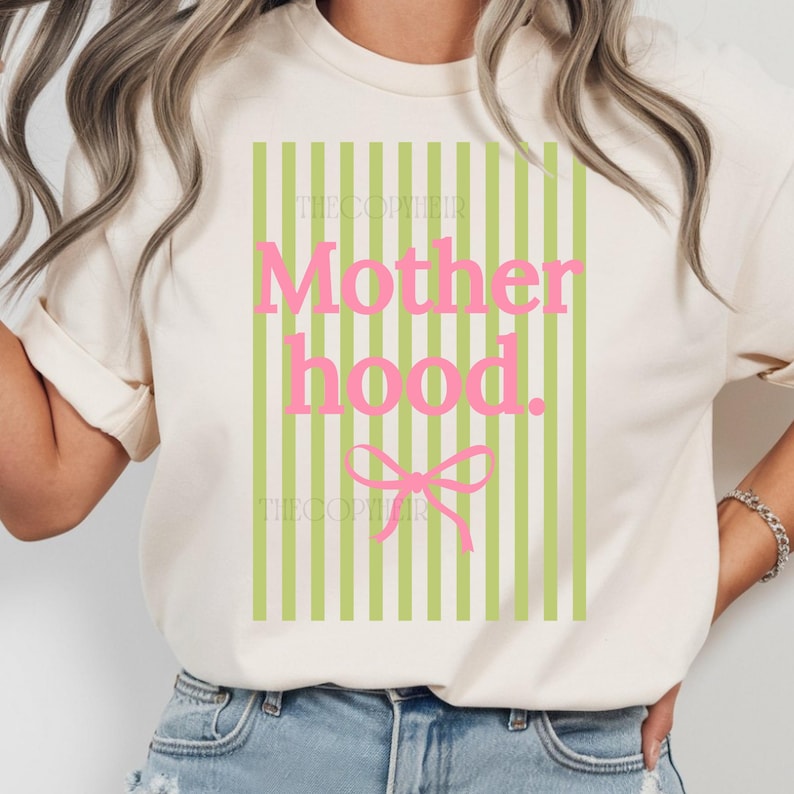 K&ouml;nnte beinhalten: Cremefarbenes T-Shirt mit rosa und gr&uuml;nem Design. Das Design zeigt das Wort "Motherhood" in Rosa, eine rosa Schleife und gr&uuml;ne vertikale Streifen. Der Text "THECOPIEIR" ist oberhalb und unterhalb des Haupttextes aufgedruckt.