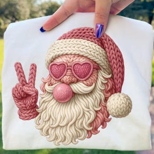 May include: White t-shirt featuring a knitted Santa Claus design. Santa wears heart-shaped sunglasses and a pink knitted hat. His beard and mustache are cream-colored. The Santa is giving the peace sign with his left hand. A pink bubble is in his mouth.