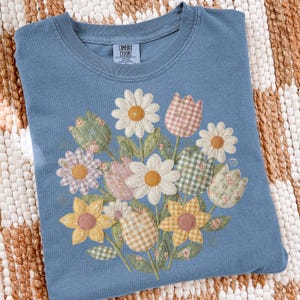 May include: A blue Comfort Colors t-shirt with a floral bouquet design. The bouquet features daisies, tulips, and sunflowers in patterns and colours like yellow, pink, and green. The shirt has a crew neck.