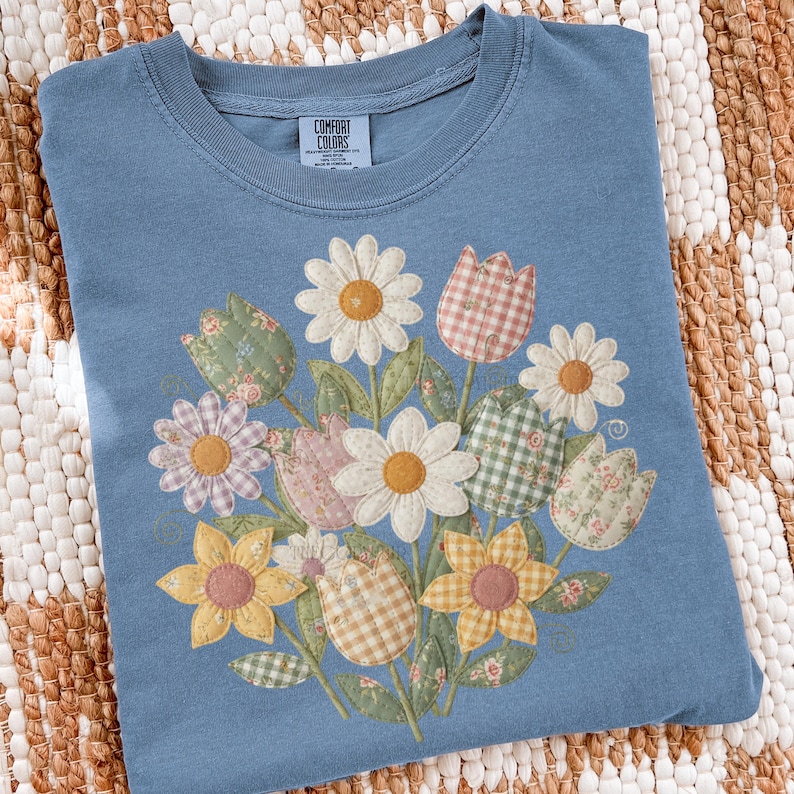 May include: A blue Comfort Colors t-shirt with a floral bouquet design. The bouquet features daisies, tulips, and sunflowers in patterns and colours like yellow, pink, and green. The shirt has a crew neck.