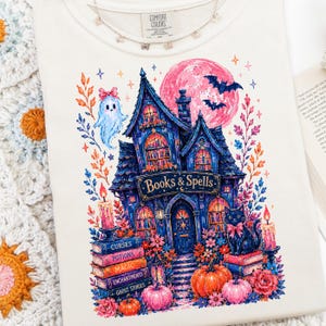 May include: A cream-colored t-shirt featuring a colorful graphic of a dark blue house labeled Books & Spells. The design includes a ghost, a black cat, pumpkins, candles, and a pink moon, surrounded by floral elements and stacks of books.