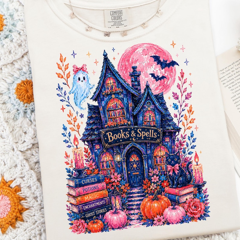 May include: A cream-colored t-shirt featuring a colorful graphic of a dark blue house labeled Books & Spells. The design includes a ghost, a black cat, pumpkins, candles, and a pink moon, surrounded by floral elements and stacks of books.