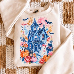 May include: A cream-colored crewneck sweatshirt featuring a colorful graphic print of a blue haunted house, ghosts with pink bows, orange pumpkins, a black cat, bats, and a pink moon surrounded by various floral elements and lit candles.