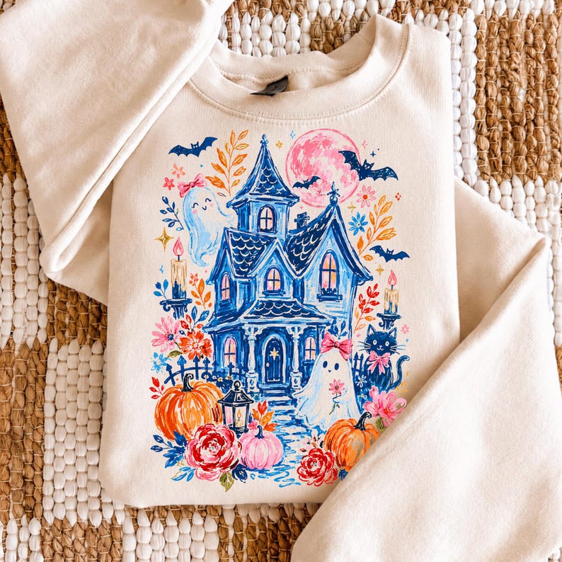 May include: A cream-colored crewneck sweatshirt featuring a colorful graphic print of a blue haunted house, ghosts with pink bows, orange pumpkins, a black cat, bats, and a pink moon surrounded by various floral elements and lit candles.