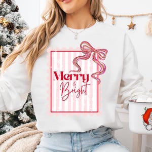 Puede incluir: Sudadera blanca con un dise&ntilde;o de rayas rosas y blancas y las palabras "Merry & Bright" en escritura roja y rosa. Un gr&aacute;fico de lazo rosa est&aacute; encima del texto. Una prenda festiva de vacaciones.
