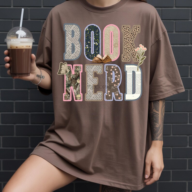 May include: Brown t-shirt with the words "BOOK NERD" in a colorful, collage-style design. The letters are filled with various patterns, including floral, leopard print, and book pages. A person is holding a beverage in a clear cup.