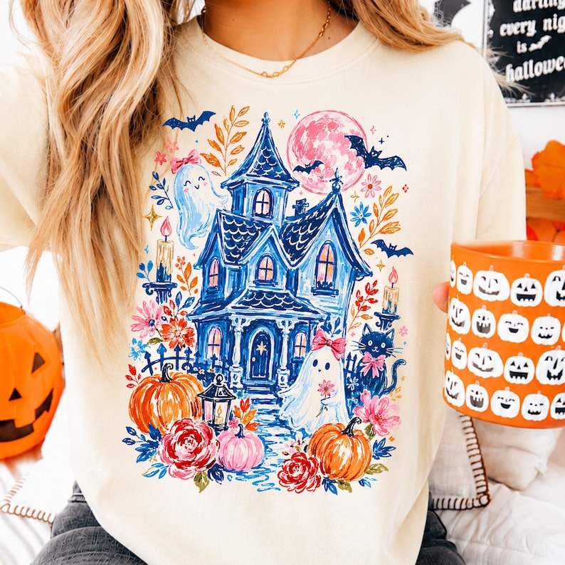 May include: A cream-colored crewneck sweatshirt featuring a colorful graphic print of a blue haunted house, ghosts with pink bows, orange pumpkins, a black cat, bats, and a pink moon. The design includes floral accents and lit candles.