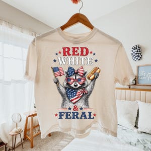 K&ouml;nnte beinhalten: Cremefarbenes T-Shirt mit einem Waschb&auml;ren, der eine Sonnenbrille, eine Schleife und ein Halstuch in Rot, Wei&szlig; und Blau tr&auml;gt. Der Waschb&auml;r h&auml;lt einen Hotdog und eine amerikanische Flagge. Der Text "RED WHITE & FERAL" ist gedruckt.