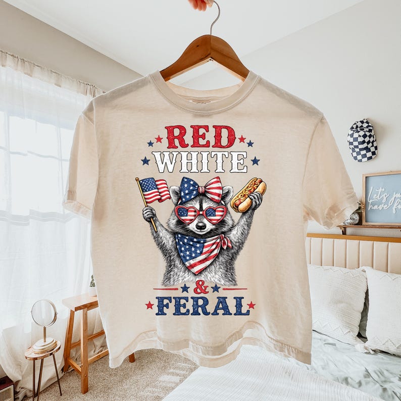 K&ouml;nnte beinhalten: Cremefarbenes T-Shirt mit einem Waschb&auml;ren, der eine Sonnenbrille, eine Schleife und ein Halstuch in Rot, Wei&szlig; und Blau tr&auml;gt. Der Waschb&auml;r h&auml;lt einen Hotdog und eine amerikanische Flagge. Der Text "RED WHITE & FERAL" ist gedruckt.