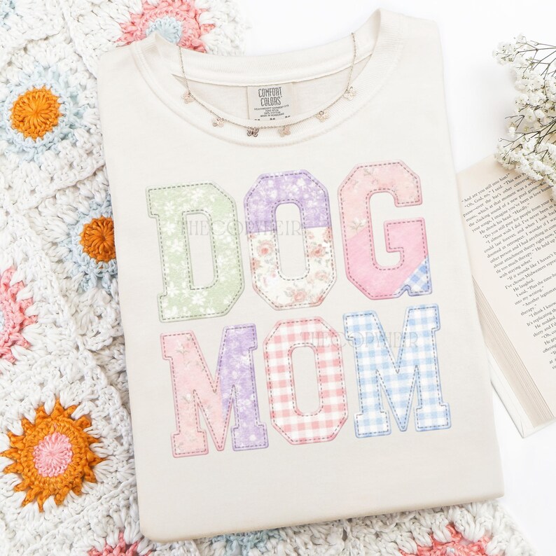 May include: Cream-colored Comfort Colors t-shirt with the words "DOG MOM" in colorful patchwork letters. The letters feature floral, gingham, and solid patterns. A delicate necklace and a crocheted blanket with floral accents are also visible.